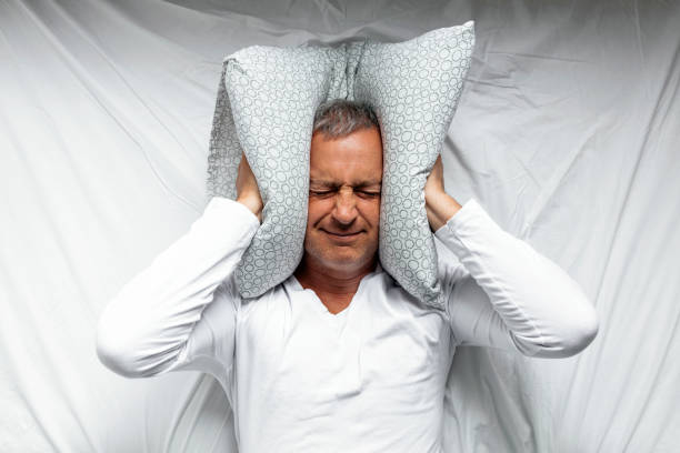 Mature man feeling uncomfortable in bed One mature man trying to sleep covering his ears to avoid neighbour noise at home or hotel during the day. hands covering ears stock pictures, royalty-free photos & images