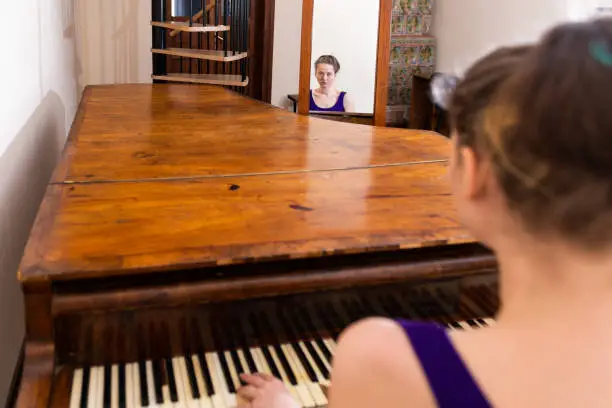 Wooden forte piano with young girl woman sitting pianist playing in formal dress in Europe for concert and mirror reflection by staircase