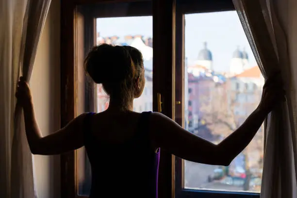 Photo of Woman back opening curtains looking from apartment room glass window with view of Lviv, Ukraine old market town square in elegant dress
