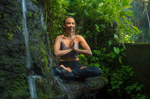 outdoors adventure portrait of young beautiful and happy woman doing yoga and meditation exercise sitting in lotus posture on rock at tropical rainforest enjoying nature during Summer holidays