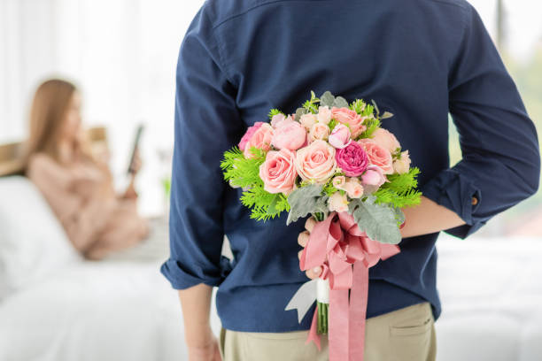 Man hiding flower at back. Man hiding buch of colorful flower at back prepare to give for woman who sitting on bed and playing smartphone in blur background. Selective focus on flower bouquet. Concept of love and anniversary. man flower stock pictures, royalty-free photos & images
