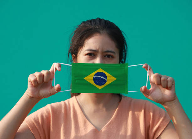 A woman with Brazil flag on hygienic mask in her hand and lifted up the front face on green background. Tiny Particle or virus corona or Covid 19 protection. stock photo