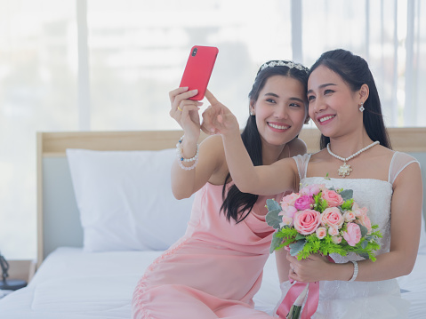 Bride and bridesmaid taking selfies on the white bed. Portrait of two young Asian women taking selfies with a smartphone in a fitting room prepare for the wedding.