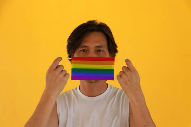 un hombre con bandera lgbt+ en máscara higiénica en la mano y levantó la cara frontal sobre fondo amarillo. diminuta partícula o virus corona o covid 19 protección. - arms lifted fotografías e imágenes de stock