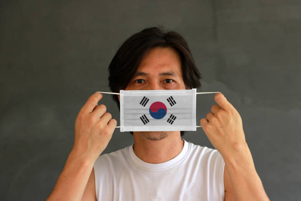 un homme avec le drapeau de corée du sud sur le masque hygiénique dans sa main et a soulevé le visage avant sur le fond gris foncé. tiny particle ou virus corona ou covid 19 protection. - arms lifted photos et images de collection