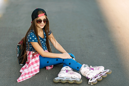 A girl in a cap and sunglasses on rollers is resting while sitting on the road, looking at the camera and smiling.