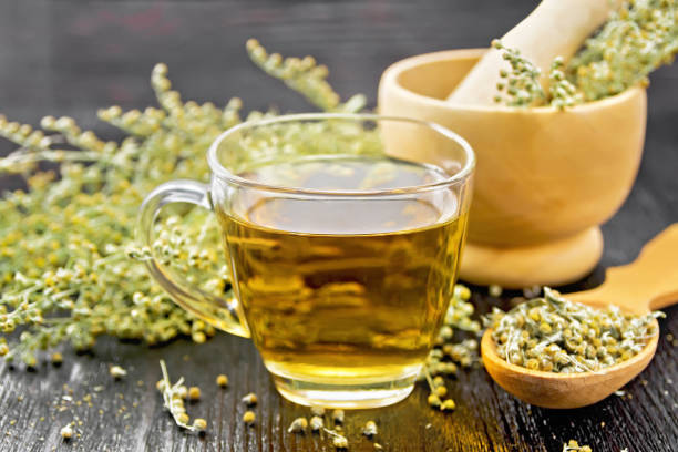 Tea of gray wormwood in glass cup with mortar on dark board Gray wormwood herbal tea in a glass cup, dry sagebrush flowers in spoon, fresh flowers in a mortar and on table against black wooden board absinthe spoon stock pictures, royalty-free photos & images