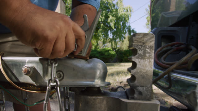 Close-Up Shot of a Hispanic Man's Hands Attach a Trailer Coupler with a Latch to the Ball Hitch on His Vehicle on a Sunny Day