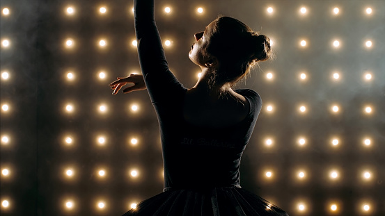 Silhouette of ballerina in black tutu is dancing ballet in the dark studio with smoke and the lights on the background.