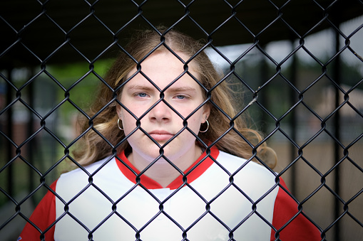 Teenage girl standing behind a fence on a baseball field.