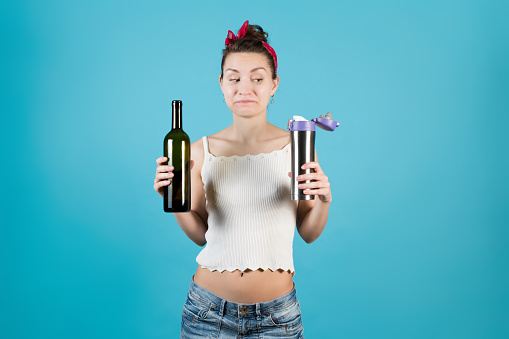 A young woman holds a wine bottle and a thermos in her hands, slyly glancing. Secretive alcoholics concept