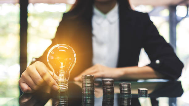 istock Businesswoman holding lightbulb next to coins stack on table 1221155619