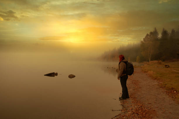 Profile view of redhead backpacker, standing on the edge of the lake, admiring the view with the fog