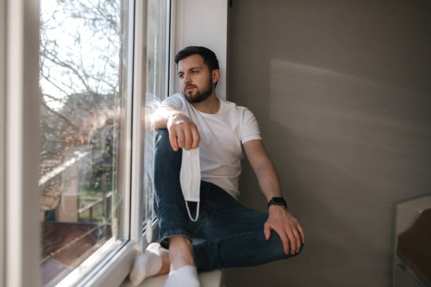 istock Young man sits on windowsill and hold in hands protective mask and looks out the window. Alone at home in quarantine. Coronavirus theme. Stay at home 1221171084