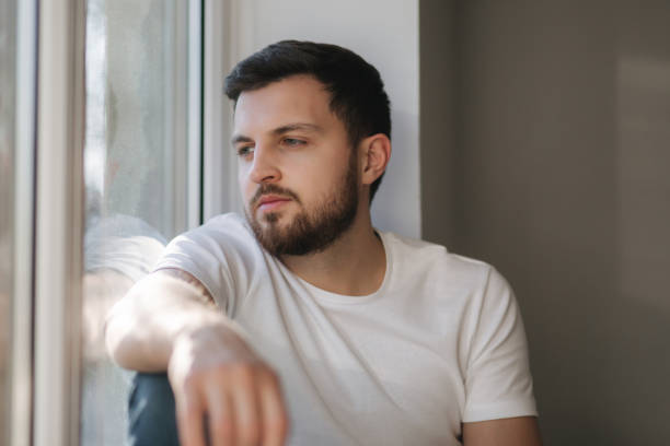 istock Young man sits on windowsill and hold in hands protective mask and looks out the window. Alone at home in quarantine. Coronavirus theme. Stay at home 1221171106