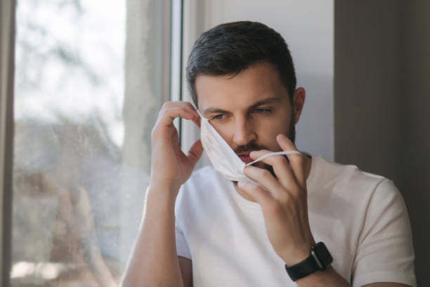 istock Young man sits on windowsill and out on protective mask and look s out the window. Alone at home in quarantine. Coronavirus theme. Stay at home 1221171143