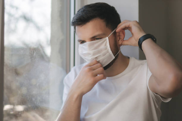 istock Young man sits on windowsill and out on protective mask and look s out the window. Alone at home in quarantine. Coronavirus theme. Stay at home 1221171676