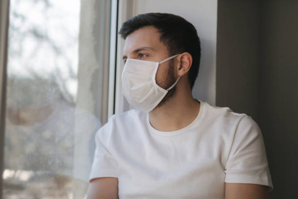 istock Young man sits on windowsill and out on protective mask and look s out the window. Alone at home in quarantine. Coronavirus theme. Stay at home 1221171684