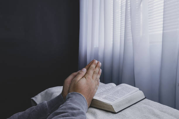 istock Man reading  Holy Bible on a white table, black wall and window  background. Stay at home. Religion concept. 1221173959