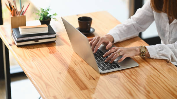 istock Cropped image of creative woman's hands typing on computer laptop that putting on wooden working desk and surrounded by stack of books, pencil holder, coffee cup and potted plant. Orderly workspace. 1221175903