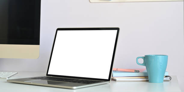 istock Photo of white blank screen computer laptop putting on white working desk surrounded by computer monitor, coffee cup and stack of books over white wall as background. 1221175952