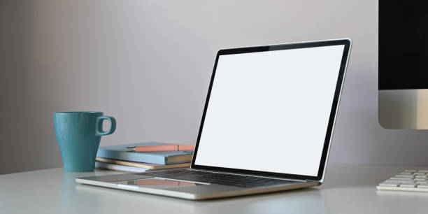 istock Photo of white blank screen computer laptop putting on white working desk surrounded by computer monitor, coffee cup and stack of books over white wall as background. 1221175994