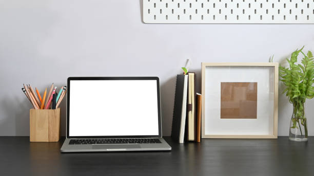 istock Laptop on working desk along with white blank screen computer, books, notebook, pencil holder, picture frame, potted plant putting together on it with white cement wall as background. 1221175999