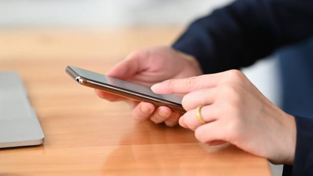 istock Photo of young woman's hands holding and using a smartphone while sitting/relaxing at the wooden working desk over comfortable living room as background. 1221176046