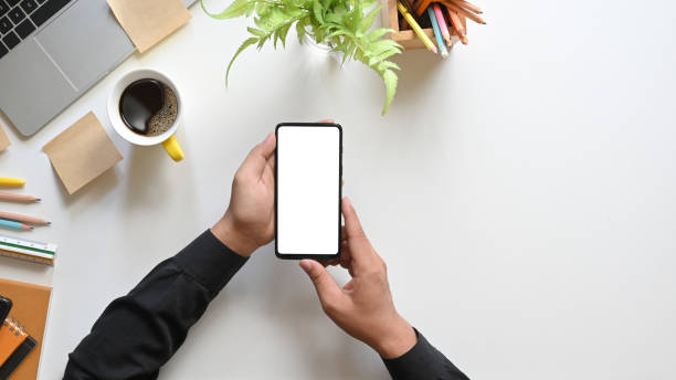 Top view hands holding a white blank screen smartphone over coffee cup, potted plant, pencils, computer laptop, notebook and ruler that putting together on white working desk.