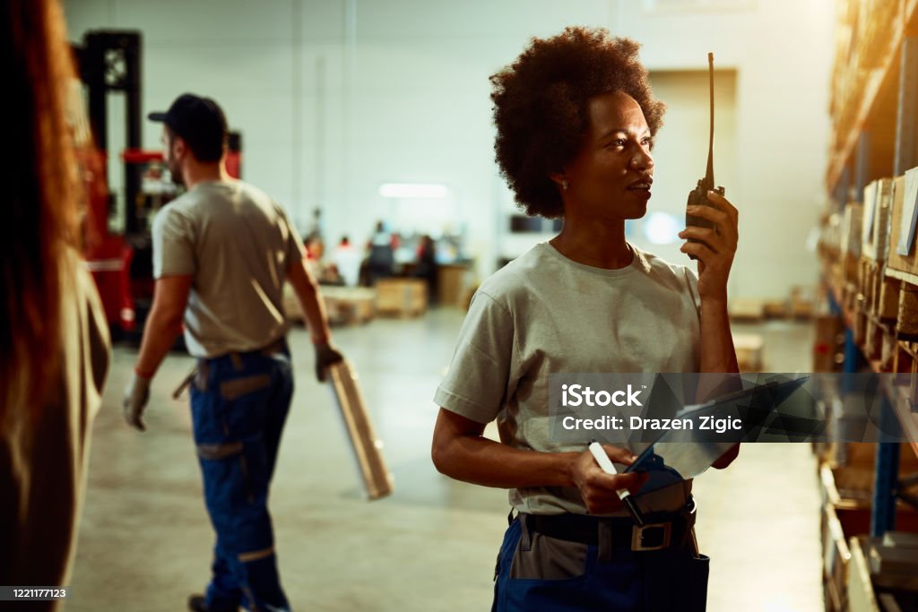African American warehouse inspector using walkie-talkie at storage compartment. Black female dispatcher communicating over walkie-talking while checking stock of products in distribution warehouse. CB Radio Stock Photo