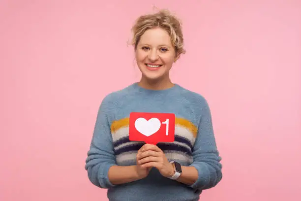 Photo of Portrait of happy adult woman with curly hair in warm sweater holding heart Like icon, love content