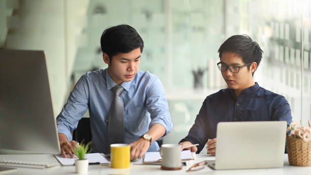 istock Photo of two smart men working together with computer laptop and tablet while sitting at the long working desk over modern office as background. 1221202197