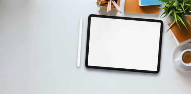 istock Top view image of computer tablet with white blank screen putting on white working desk and surrounded by stylus pen, potted plant, notebook, diary, coffee cup, pencils and pencil holder. 1221202259