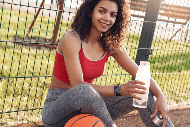 Basketball Player. Woman sitting with ball drinking water on court resting smiling happy close-up