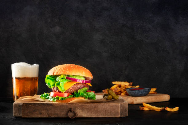 burger and beer. hamburger with beef, cheese, onion, tomato, and green salad, a side view on a dark background with pickles and french fries, with a place for text. selective focus - salad food and drink food lettuce imagens e fotografias de stock