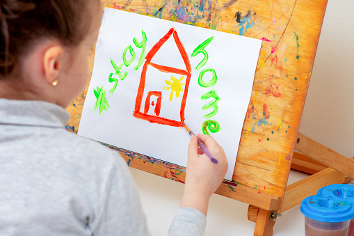 Child is drawing red house by watercolors with a written phrase over the house Stay Home on the easel. Stay Home concept.