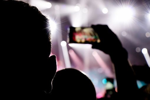 Silhouettes of a mans ear and another mans hand shooting a concert with cellphone with light effects in the background, from behind