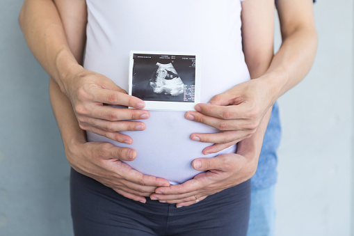 In a close-up shot, a pregnant woman at home holds an ultrasound photo and a hand-sewn symbol representing her child's zodiac sign. She gently caresses her baby bump, expressing her anticipation for the new life and the joy of soon becoming a mother.