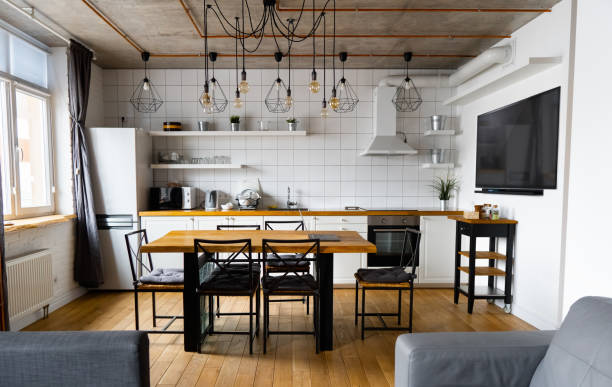 istock Modern scandinavian an eat-in kitchen interior design with big wooden table and chairs against light wood floor, bright white walls and furnitures with TV, appliances and hanging light bulbs 1221223071
