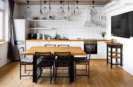 Moderno escandinavo un diseño interior de cocina comedor con gran mesa de madera y sillas contra suelo de madera clara, paredes blancas brillantes y muebles con TV, electrodomésticos y bombillas colgantes photo