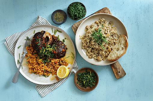 Traditional Italian Osso Buco with Creamy Saffron Risotto dish topped with fresh parsley, garlic and lemon zest and Risotto with Wild Mushrooms. Flat lay top-down composition on blue background