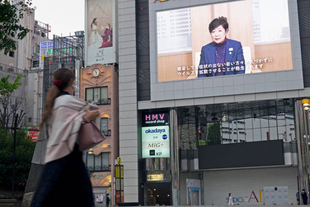 A japanese woman looks at a screen showing Tokyo's Governor Yuriko Koike's announcement Shinjuku, Tokyo, Japan - April 26, 2020: Shinjuku City became empty after state-of-emergency declaration due to COVID-19 crisis. A woman looks at a big screen showing Tokyo's Governor Yuriko Koike's announcement about COVID-19 (Coronavirus) state of emergency stock pictures, royalty-free photos & images