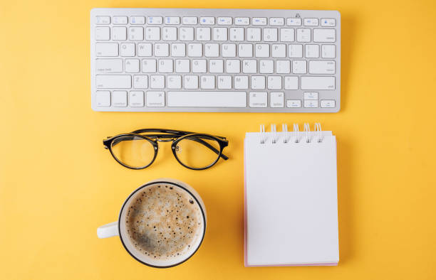 istock Home office workplace: cup of coffee, empty notebook, keyboard, tulip flowers on yellow background. Flat lay, top view. Work from home 1221238245