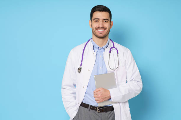 istock Medic in white robe, looking straight at camera with positive smile, holding tablet computer next to chest while working in hospital, isolated on blue background 1221257242