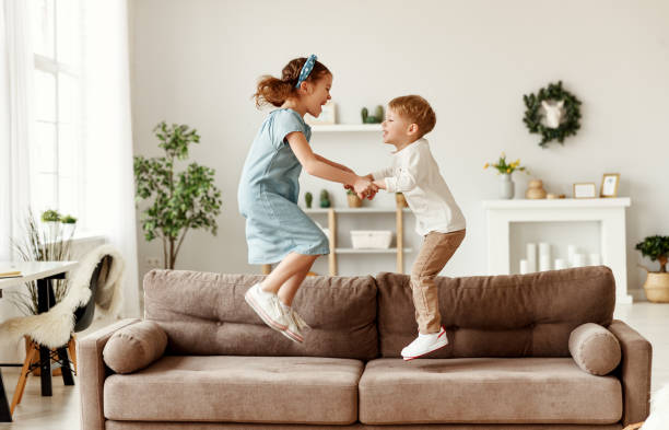 Happy siblings jumping on sofa Side view of cheerful boy and girl holding hands and jumping on couch while having fun at home together sibling stock pictures, royalty-free photos & images