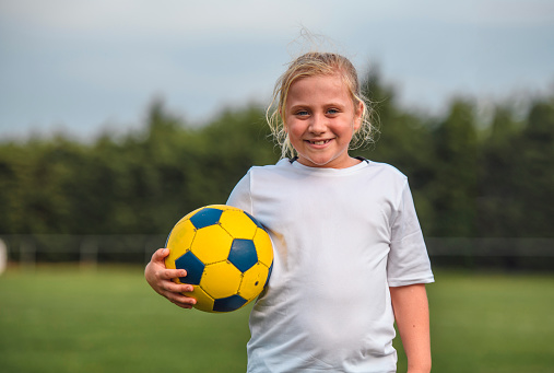 Front view of young blonde girl footballer holding ball under her arm and smiling at camera.