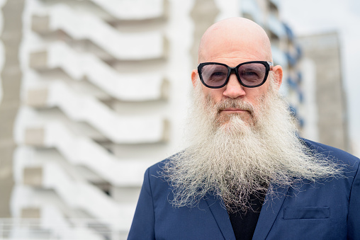 Portrait of mature handsome bald hipster businessman with long beard against view of the city