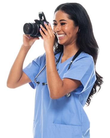 Side view of aged 20-29 years old who is beautiful with long hair latin american and hispanic ethnicity young women photographer standing in front of white background who is cheerful who is photographing and holding camera