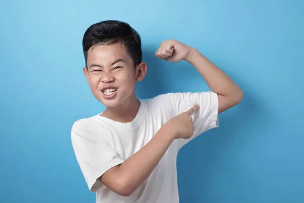 Portrait of funny strong healthy Asian boy shows his muscle and strength, confident kid shows his biceps arm muscle
