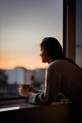 Young beautiful woman day dreaming while looking through her apartment window during coffee time. Copy space.